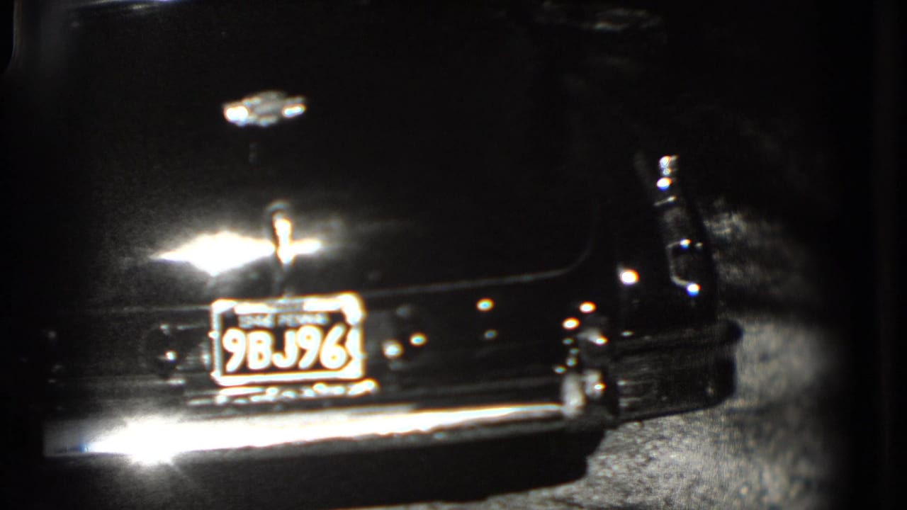 1975 Washington, DC home-movie frame showing the rear of a dark classic car with plate 9BJ96 and a reflective chrome bumper.