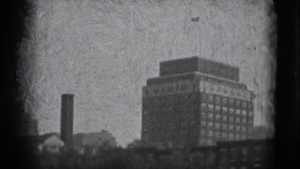 1975 Washington DC home-movie frame showing a hazy tall building with rows of windows, a nearby chimney, rooftops, and heavy 8mm grain.