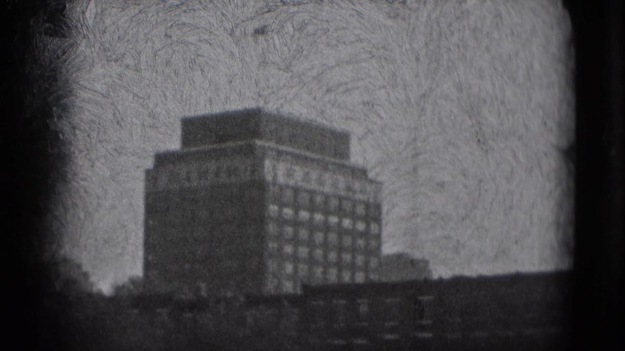 1975 Washington DC frame with a prominent rectangular building, rows of windows, low foreground roofs, and textured black-and-white 8mm film grain.