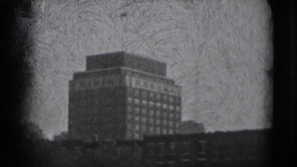 1975 Washington DC frame with a prominent rectangular building, rows of windows, low foreground roofs, and textured black-and-white 8mm film grain.