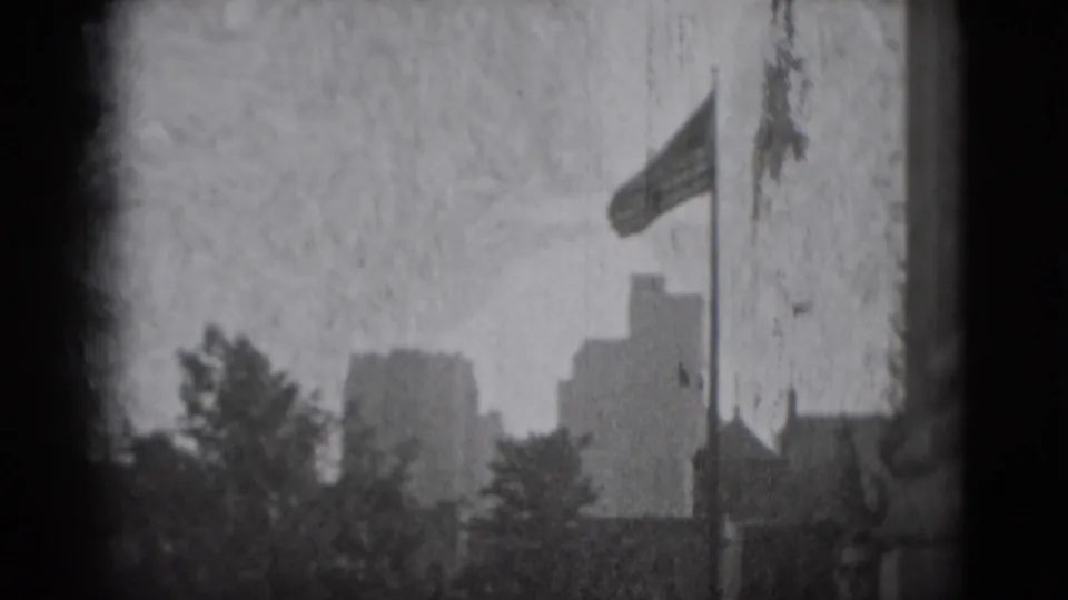 Grainy 1975 Washington DC frame showing distant blocky buildings, trees, and a flagpole against a pale sky.