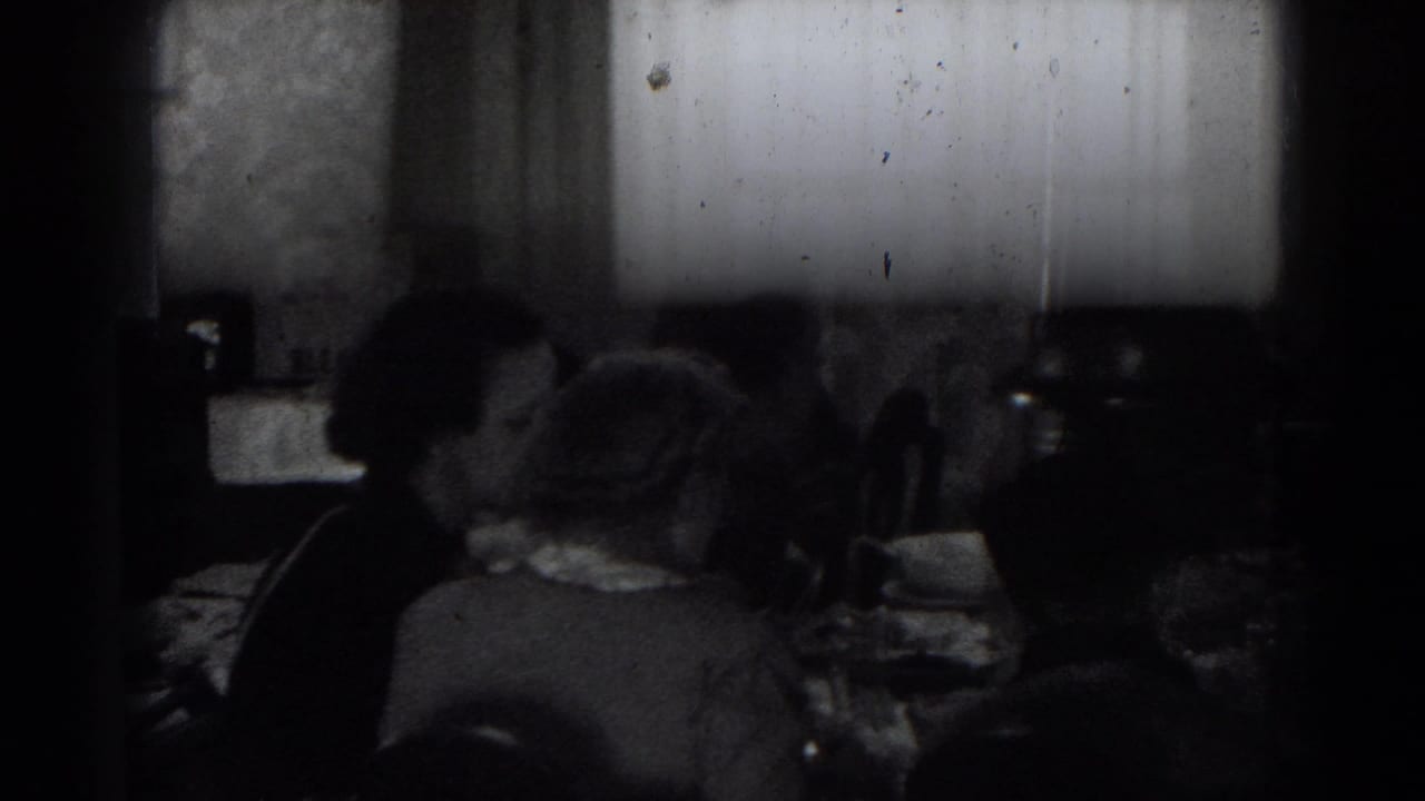 Adults sit around a dim dining table in archival home-movie footage from Lincoln, Nebraska, with plates and glassware in front of them.