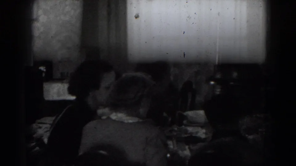Adults sit around a dim dining table in archival home-movie footage from Lincoln, Nebraska, with plates and glassware in front of them.