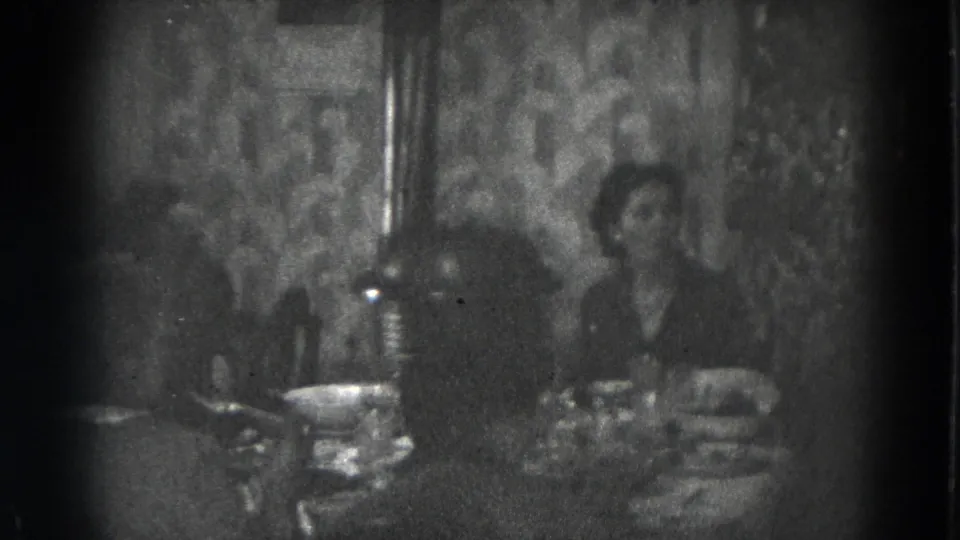 Two men sit at a dining table in a dim 8mm home-movie frame, with dishes and patterned wallpaper visible behind them.