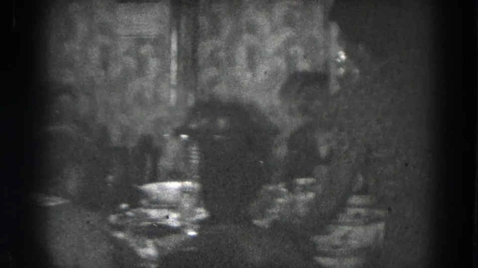 Women appear seated for a meal in a dim Lincoln, Nebraska private interior, recorded on black-and-white 8mm home-movie film.
