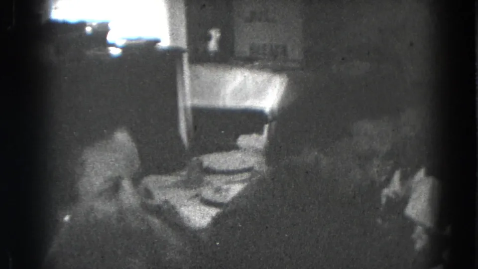 Two women appear in conversation beside a set dining table in a private Lincoln, Nebraska interior on black-and-white 8mm film.