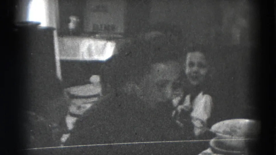 Dim black-and-white 8mm frame of seated people at a dining table in a private Lincoln, Nebraska interior.