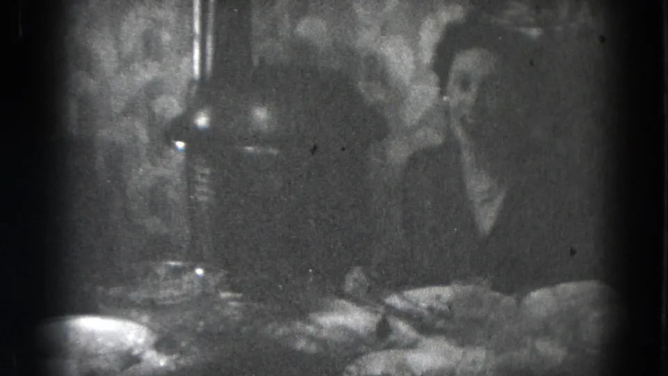 Black-and-white home-movie frame showing adults seated around a crowded dining table under hanging light fixtures.