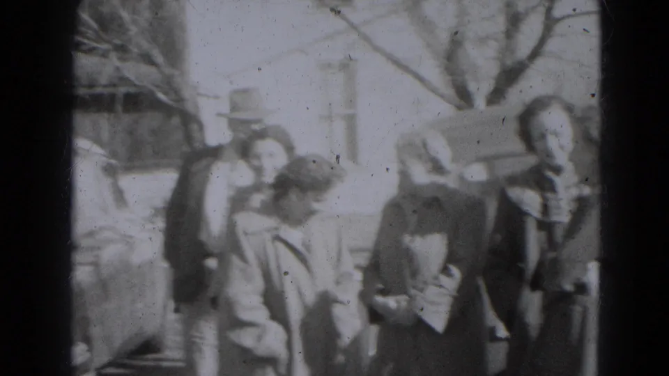 Adults in mid-century coats gather near a car and yard, with one person bending forward in the 1947 Lincoln home-movie scene.