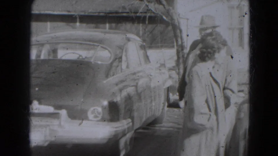 A man in a brimmed hat and a woman in a coat stand near a rounded parked car in the 1947 Lincoln, Nebraska yard scene.