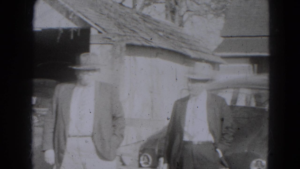 Adults in coats and hats stand near an older car outside a rustic building.
