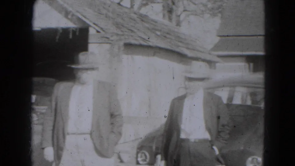 In Lincoln, Nebraska home-movie footage, formally dressed adults stand near an older car outside a rustic building.