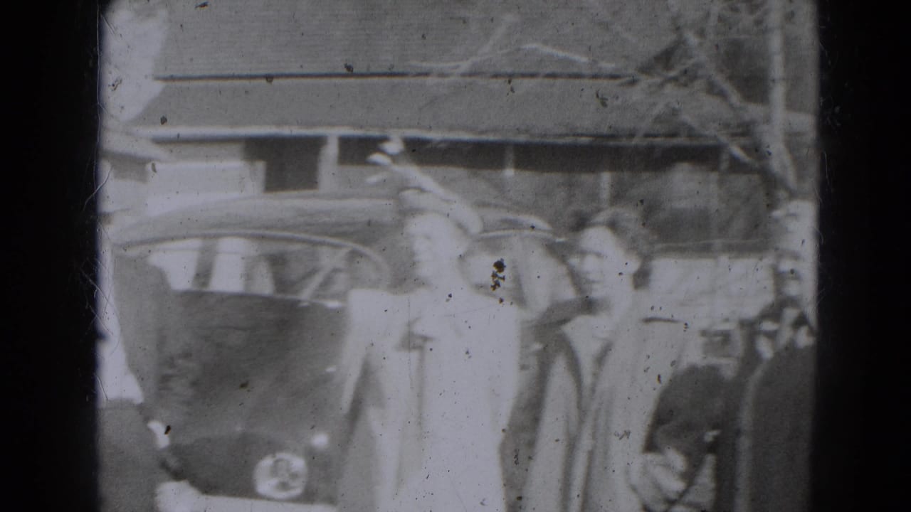 Several adults stand beside an older car, with a building and trees behind them.
