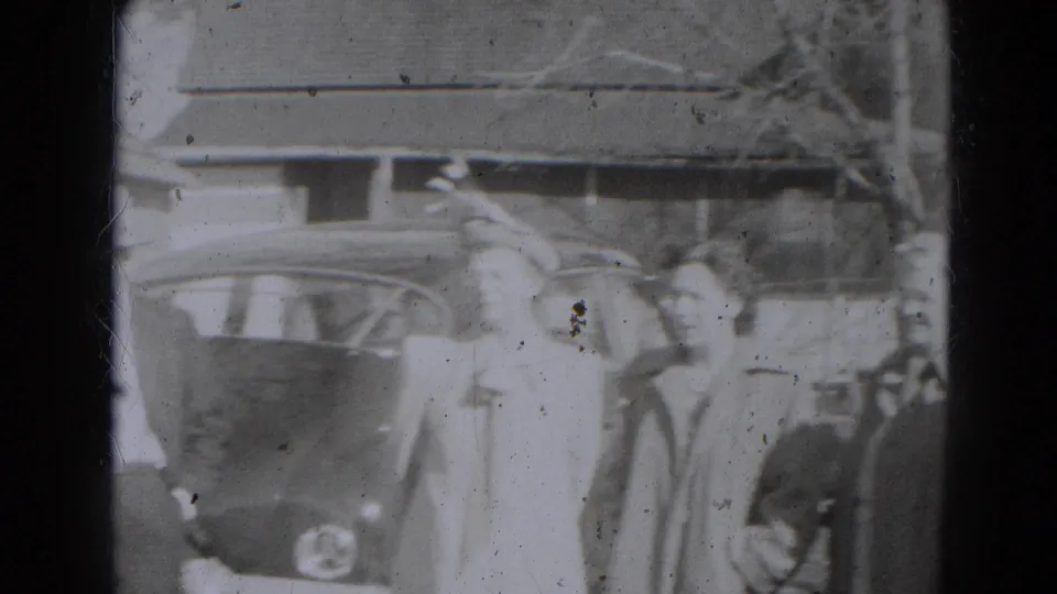 Adults in formal outerwear stand near a mid-century car in the Lincoln, Nebraska outdoor gathering scene.