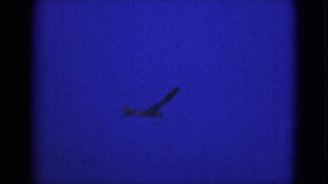 A small airplane appears as a dark silhouette against a saturated blue sky in archival 8mm footage.