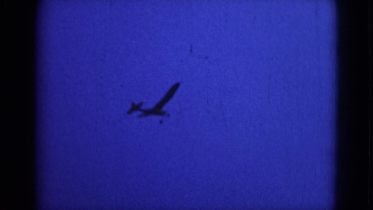 A small aircraft is seen in silhouette, angled upward against a blue sky in the Middletown home-movie footage.