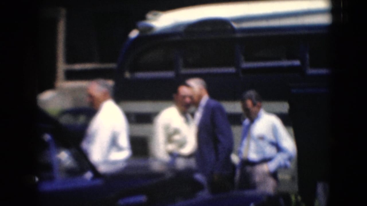 Several adult men in shirts and suits stand beside a blue-and-white bus, with one man in a light hat near the group.