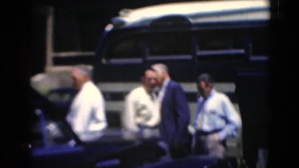 Several adult men in shirts and suits stand beside a blue-and-white bus, with one man in a light hat near the group.