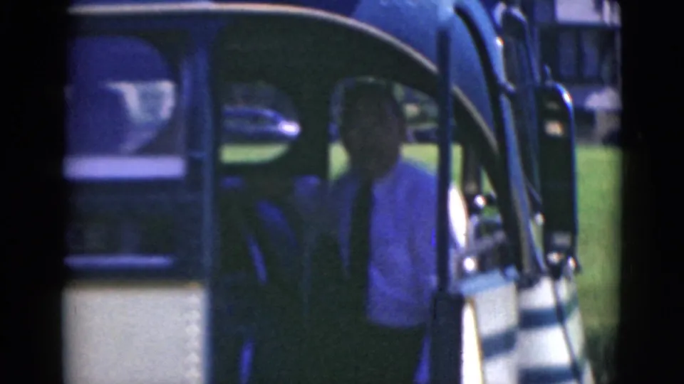 An adult man in a light jacket stands at the open door of the blue-and-white bus in grainy 8mm home-movie footage.