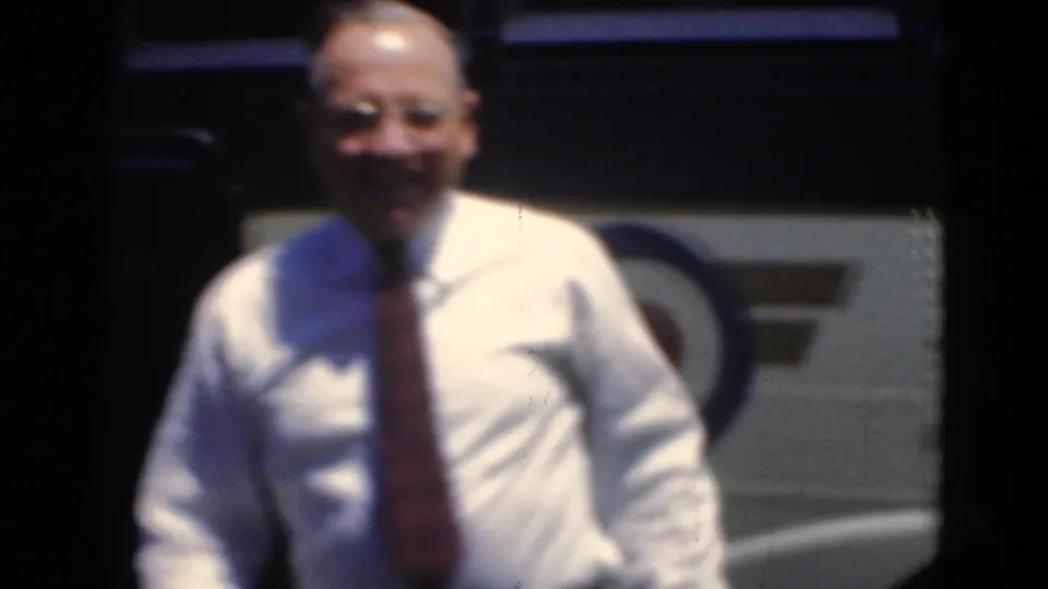 A smiling man in a light shirt and tie walks toward the camera with the bus body visible behind him.