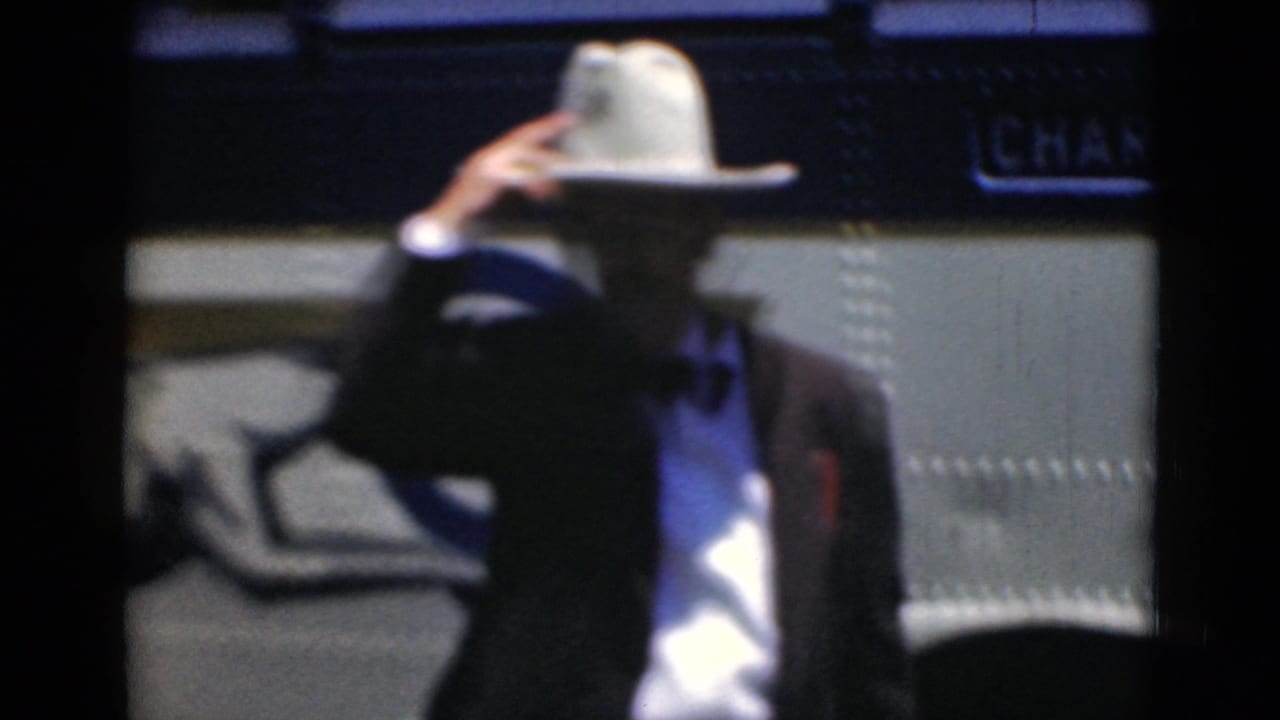 The man adjusts or tips his white hat in front of a charter bus during the Middletown, Pennsylvania home-movie scene.