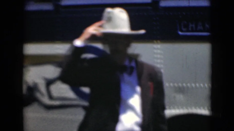 The man adjusts or tips his white hat in front of a charter bus during the Middletown, Pennsylvania home-movie scene.
