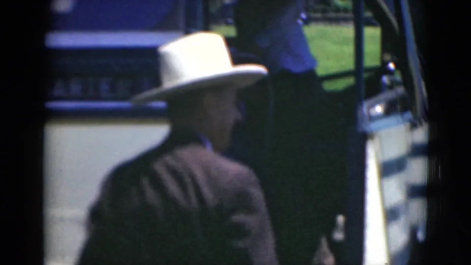A suited man in a white hat approaches or steps into an open bus doorway, with lawn visible beyond the vehicle.