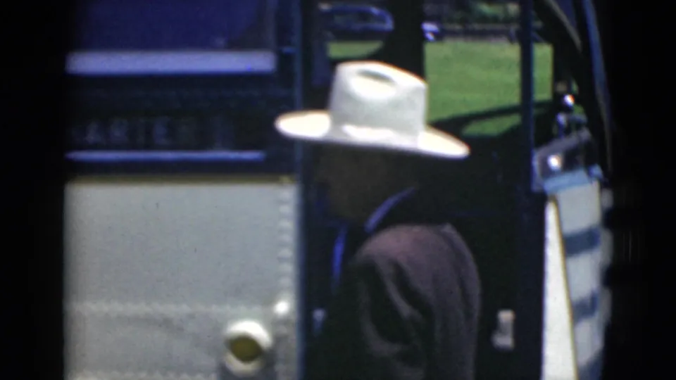 The man stands near the open charter bus entrance, partly turned away, as the home-movie camera follows his boarding.