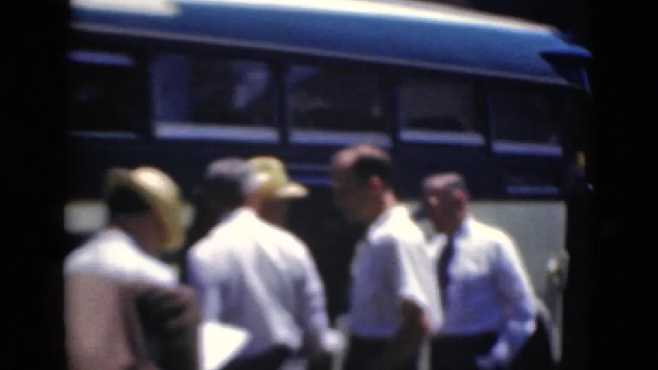 A group of adult men stands beside a bus, with white shirts, hats, and formal clothing visible in this 1947 home-movie frame.