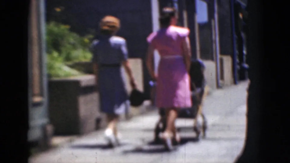 Two women in dresses walk away along a Middletown, Pennsylvania sidewalk, with one pushing a stroller in 8mm home-movie footage.