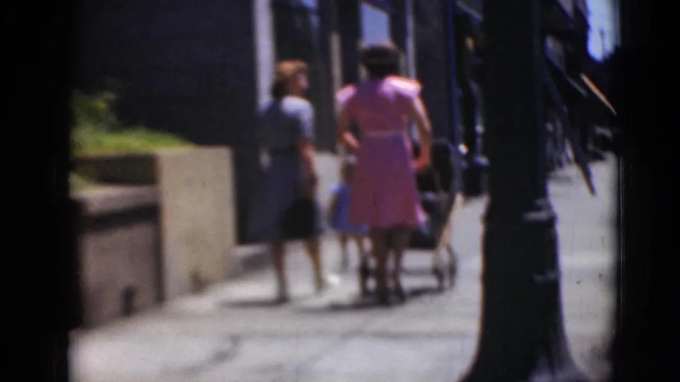 Two women in dresses are seen from behind as they walk with a stroller along a Middletown sidewalk, partly obscured by foreground framing.