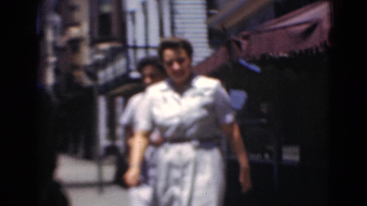 Two women walk toward the camera on a sunlit sidewalk beside storefront awnings.