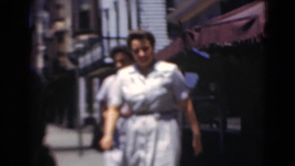 In Middletown, Pennsylvania, two women in light dresses walk side by side past storefronts on a sunlit 1940s sidewalk.