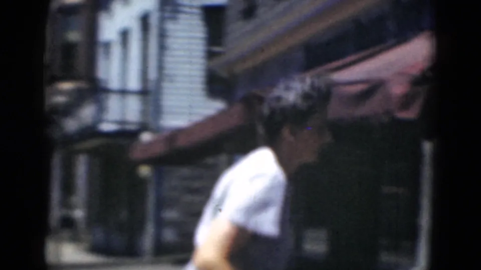 A woman in profile continues along the Middletown sidewalk, passing awnings and building fronts in soft 8mm daylight.