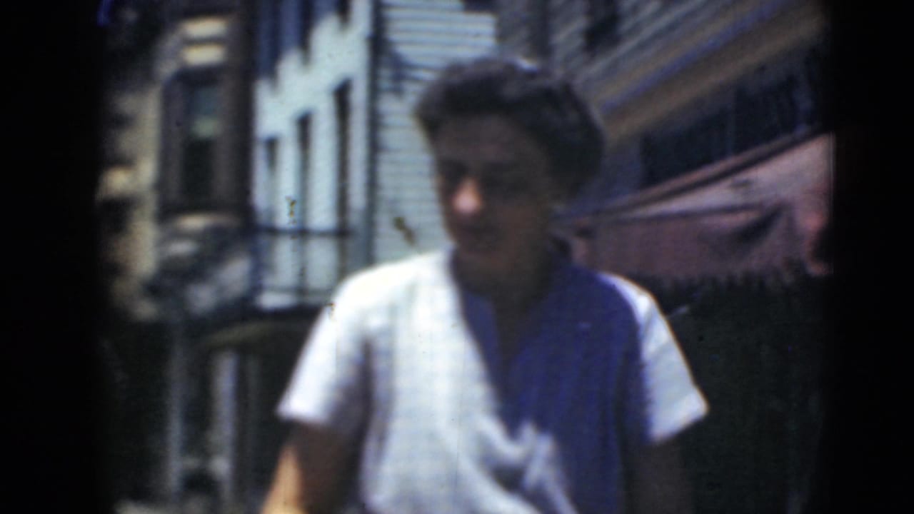 A woman in a white shirt walks through a Middletown residential block, with row houses and awnings visible in soft 8mm focus.