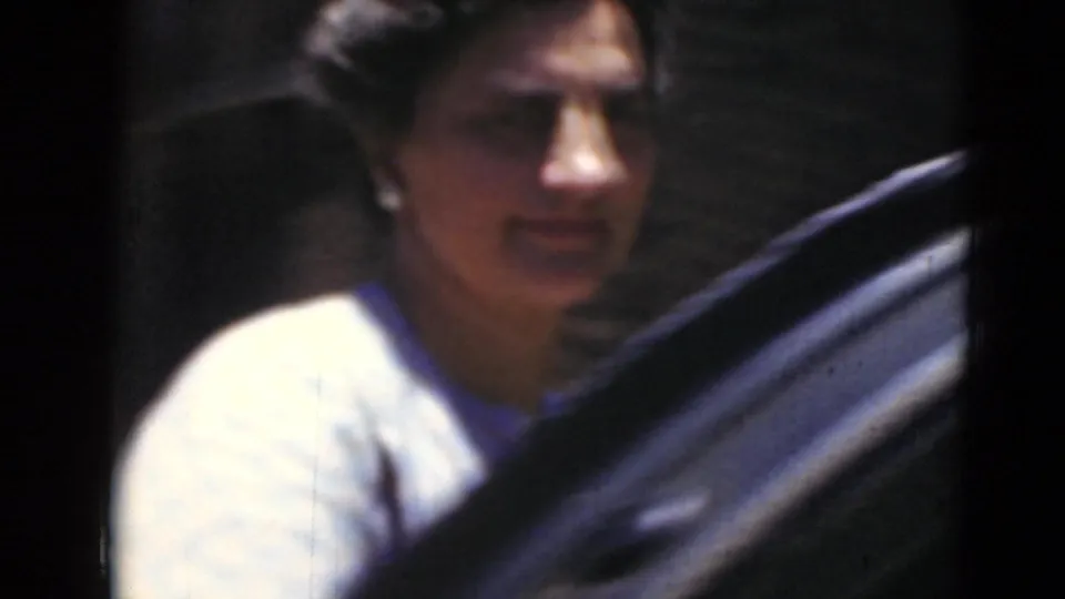 Closer 8mm frame of the woman near a dark car, her face turned toward the camera as she appears to smile or speak.