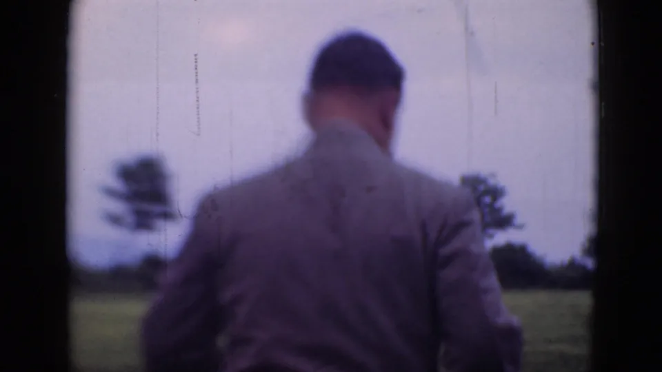 An adult man in a suit is seen from behind walking away across an open grassy area, with trees and sky in soft 8mm focus.