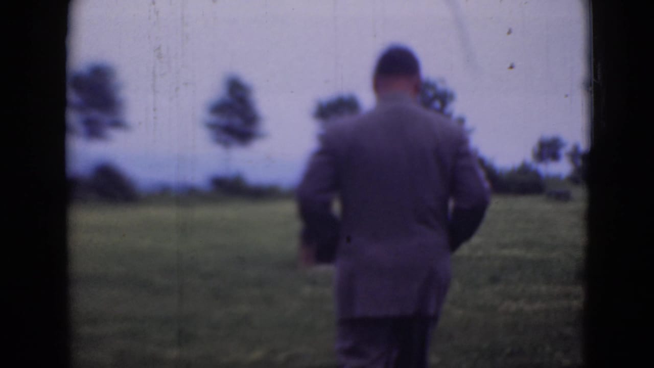 Man in a light suit walks away across grass with trees along the horizon.