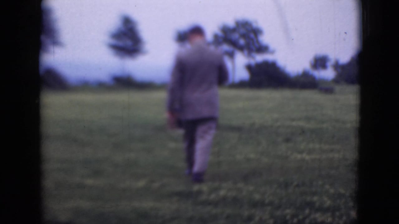 A suited man walks away across a grassy field under an overcast sky.