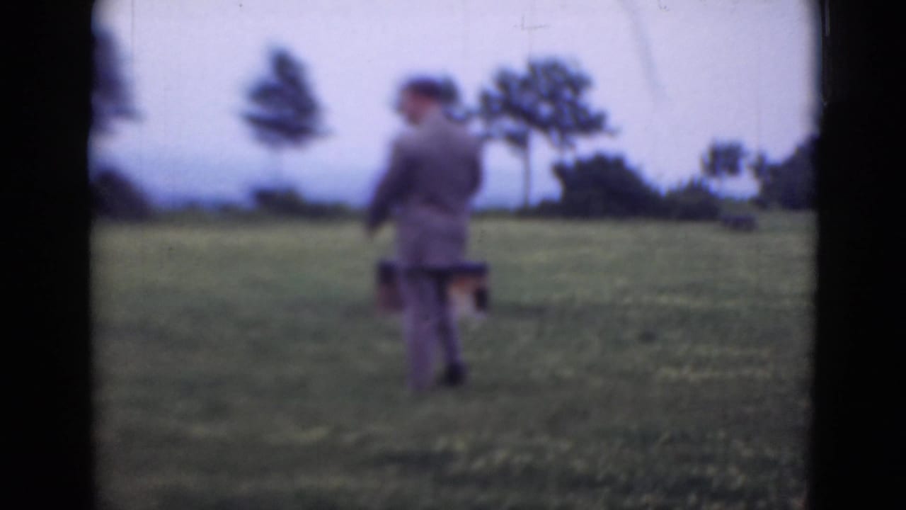 A suited man carries a dark case while walking through a grassy field.