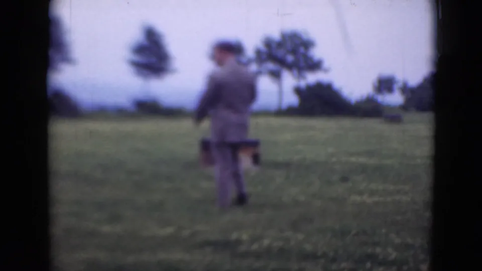 The 1947 Middletown home-movie frame shows a man in a suit and hat walking through grass while holding a dark case.