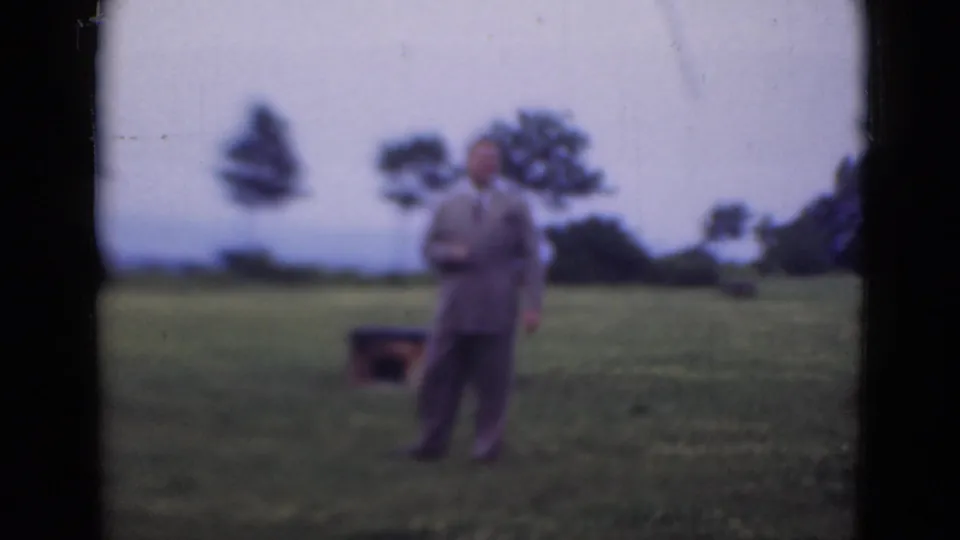 In the Middletown, Pennsylvania 1947 footage, the suited man turns toward the camera beside a dark case on the grass.