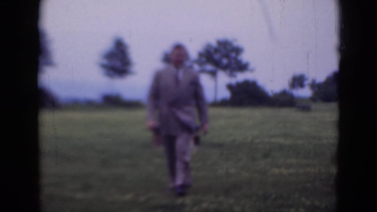 Adult man in a light suit walking toward the camera across a grassy field.