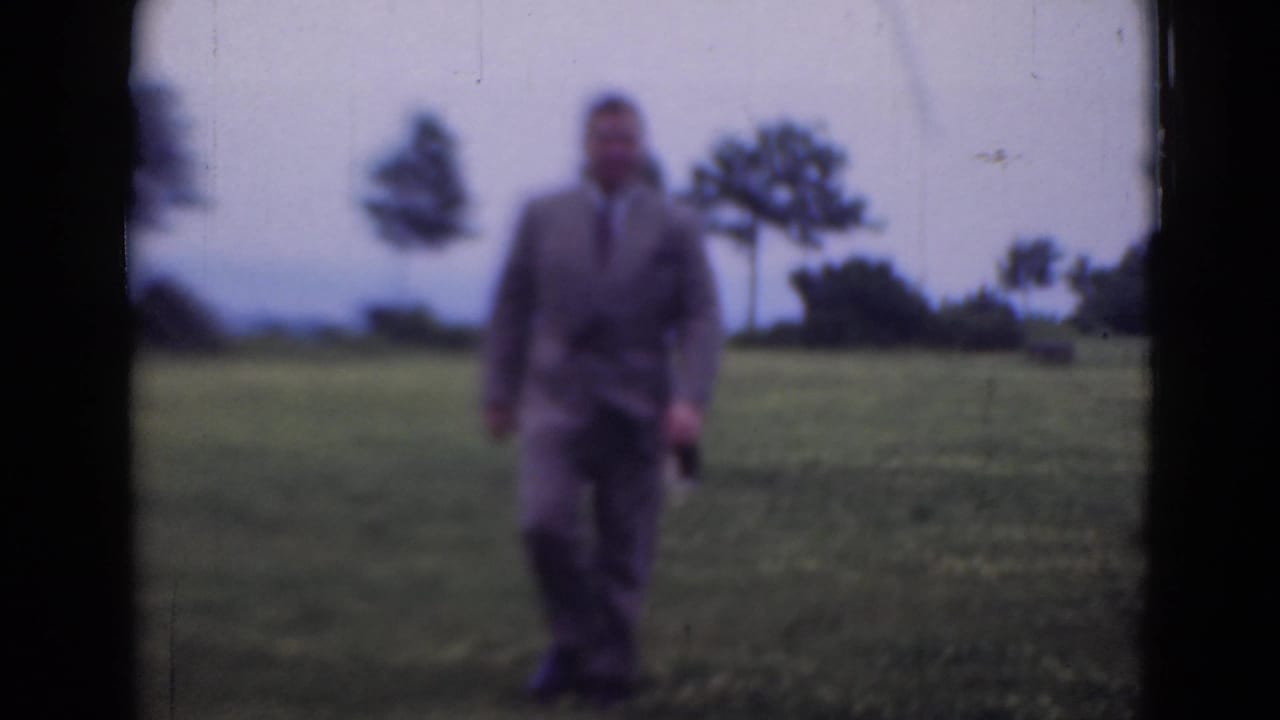 The man remains in the open field, framed softly against grass, trees, and a pale sky in 1947 home-movie footage.