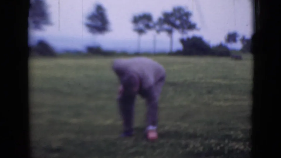 The suited man bends toward the ground in the grassy field, suggesting he is inspecting or searching the area.