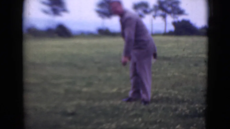 The suited man stands in profile in the field, with soft-focus trees and open grass behind him.