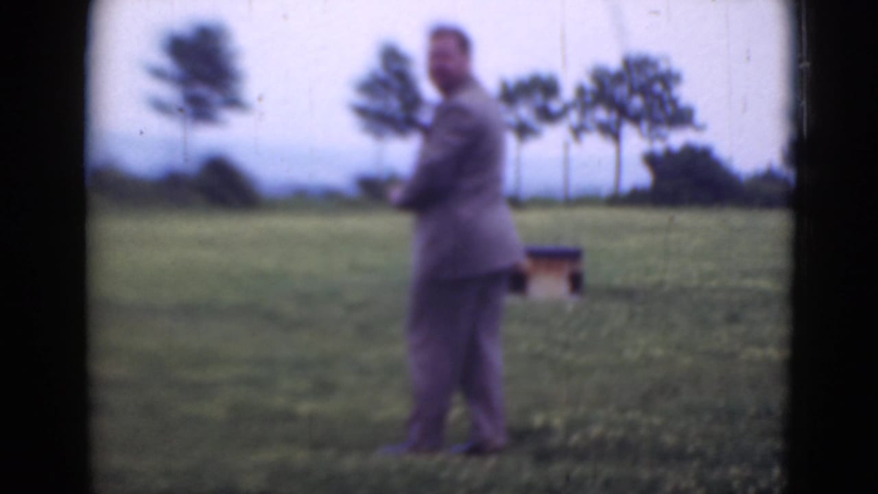 Man in a light suit stands in a grassy field with trees and a small structure behind him.