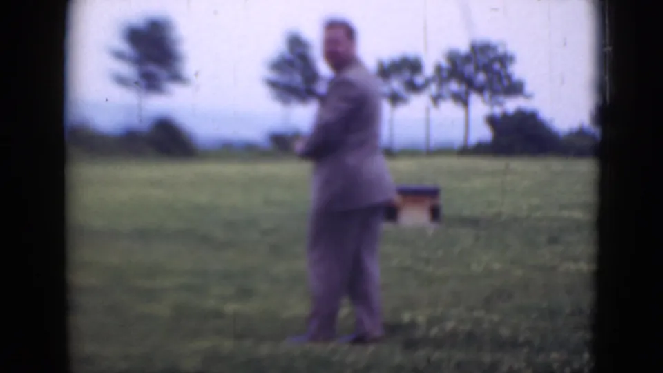 In Middletown, Pennsylvania home-movie footage, a suited man stands in an open grassy field with distant trees and a small wooden structure.