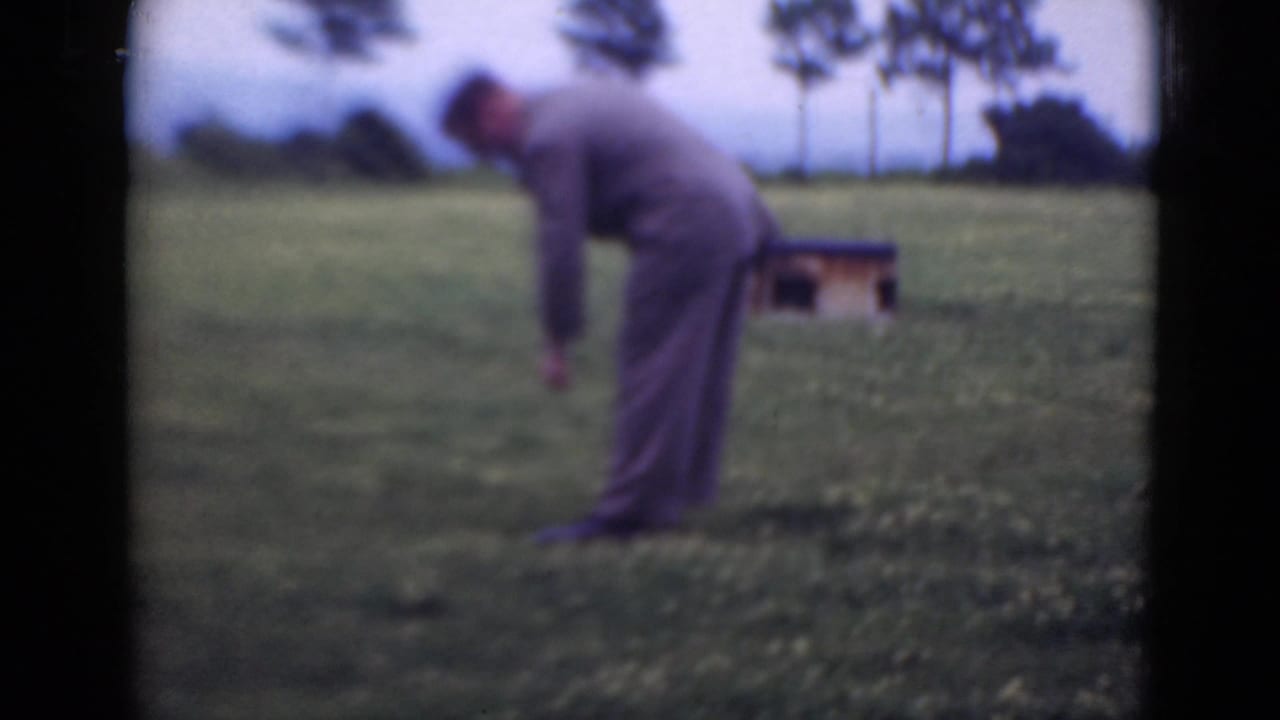 The man bends down in the grassy field, with a small wooden structure and a line of trees visible behind him.