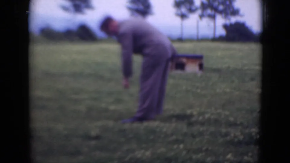 The man bends down in the grassy field, with a small wooden structure and a line of trees visible behind him.